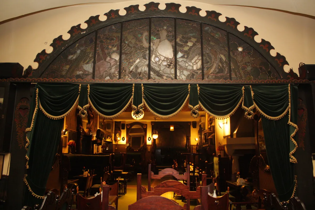 A carved arch with a band of stained glass above dark green velvet drapes, framing a warmly lit room with wooden chairs.