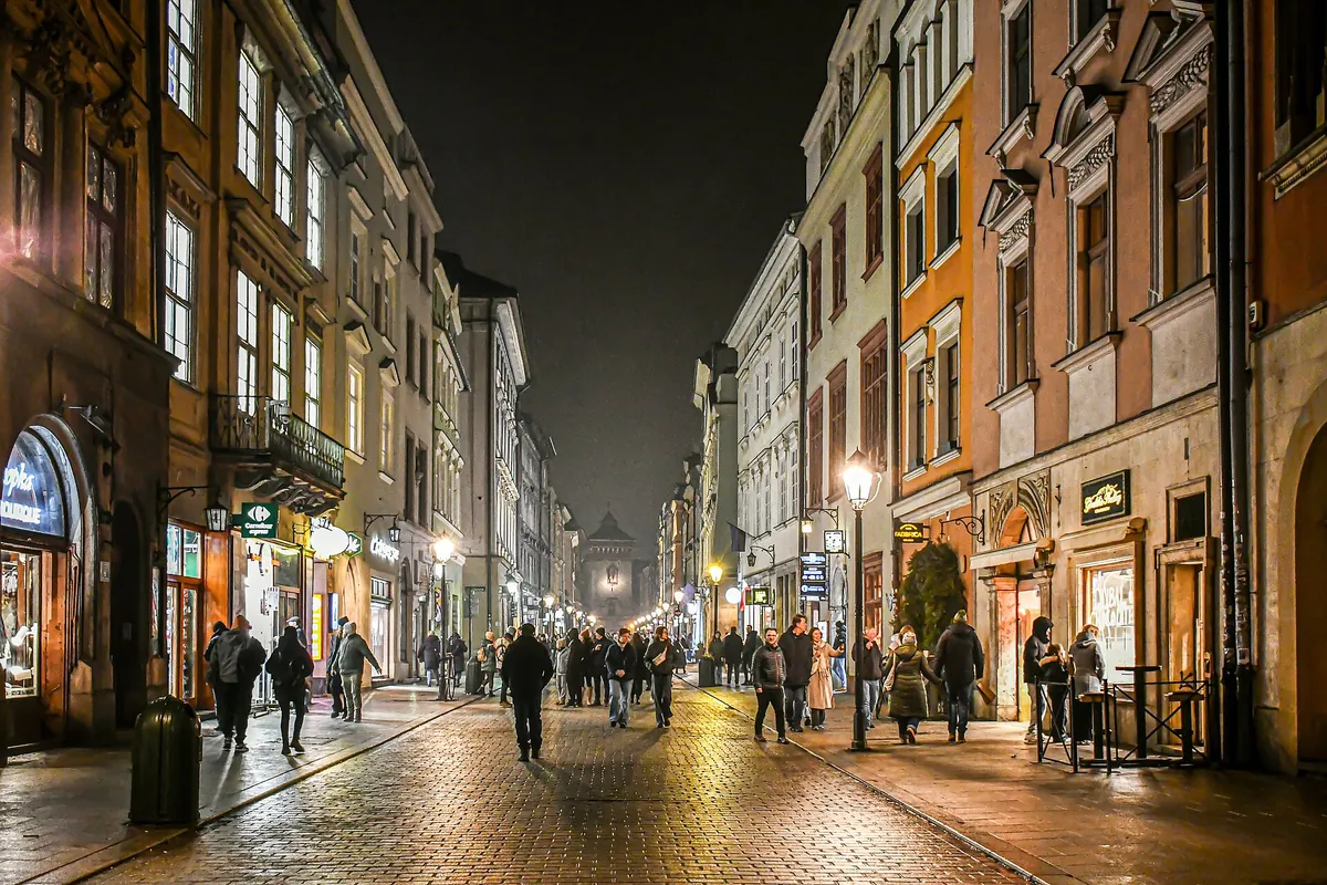 Floriańska Street at night, a cobbled street lit by lamps and shop windows, with people walking and the city gate at the far end.