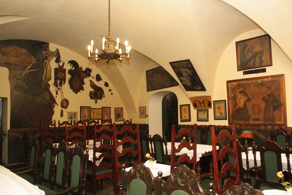 A vaulted room with a chandelier, framed caricatures and paintings on cream walls, and dark wooden chairs with green seats.